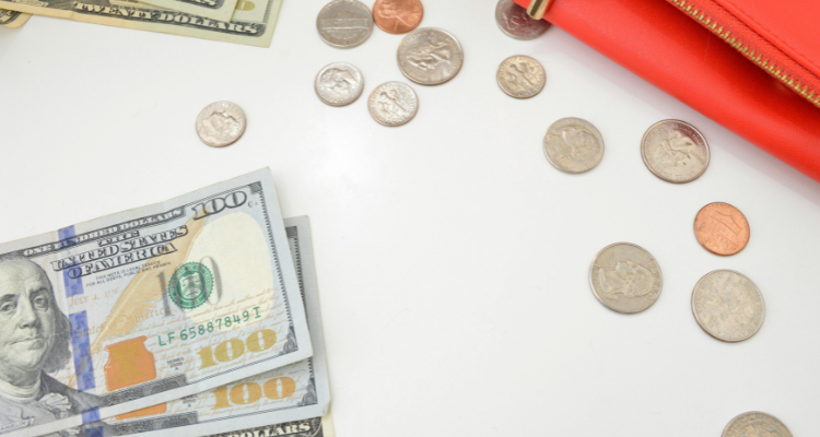 Scattered U.S. currency with coins and bills beside a red wallet on a white surface