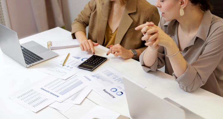 Two women working in accounting & bookkeeping with laptops, documents, and a calculator at a modern office desk.